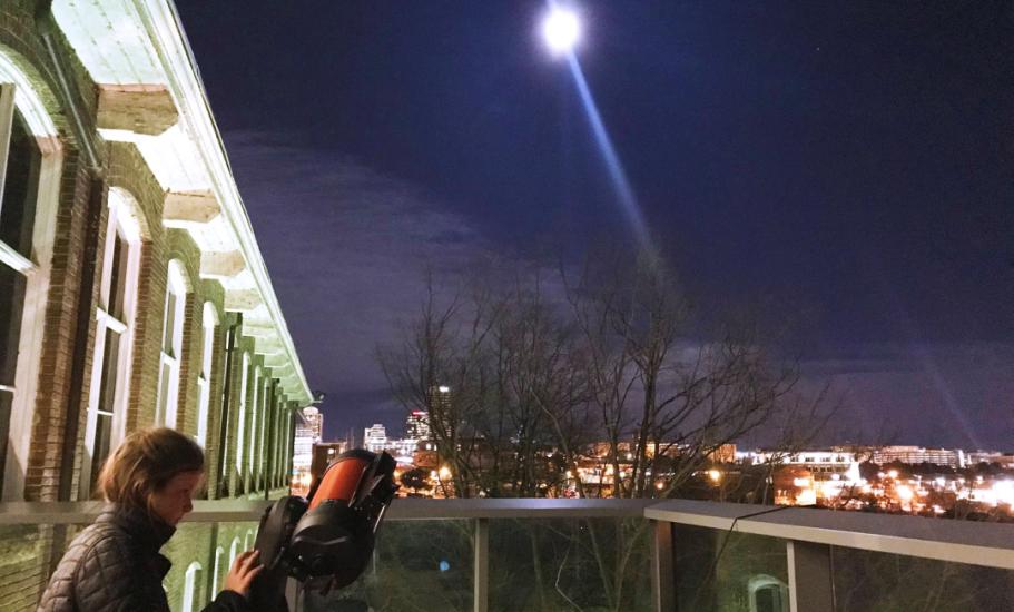 Woman stands makes adjustment to telescope pointed at Moon which is visible in the night sky from the Observatory Terrace. Columbia skyline visible in background.