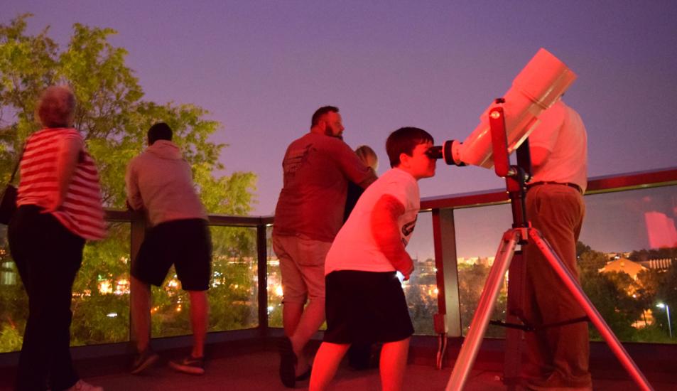 Group of people stand on terrace in the early evening with one young boy looking at the night sky through binoculars in the foreground