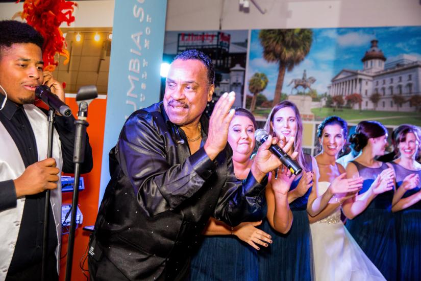 Two men perform with one singing and one about to clap as wedding party stands with them and claps along