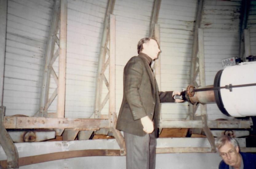 Man in brown suit jacket stands with hand on the eye piece of a large white telescope
