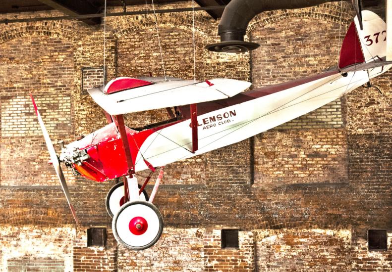 White biplane with red trim and nose that reads 'Clemson' on the side hangs from ceiling on display at the State Museum