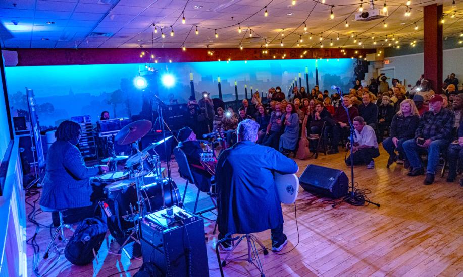Large room lit by string lights and blue spotlights shows drummer and two guitarists performing for seated audience