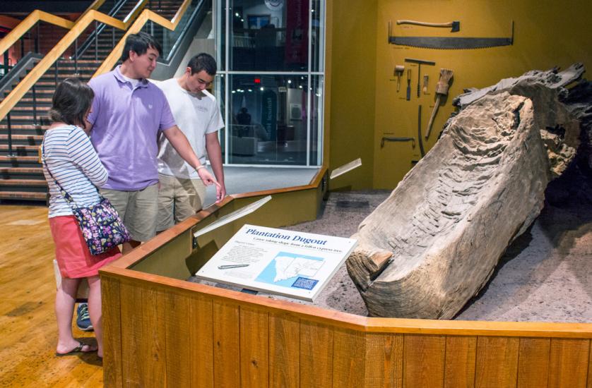 Three adults stand to the left of a display of a large hollowed out canoe on exhibition