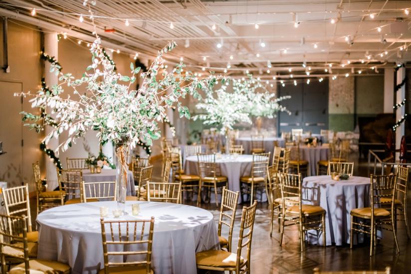 Large space with string lights suspended from ceiling and round tables with silver table cloths, gold chairs and large flower center pieces.