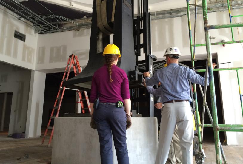 Two museum staff members, a man and a woman, in hard hats stand with back to camera facing a large black metal base being lowered down onto a concrete pier.