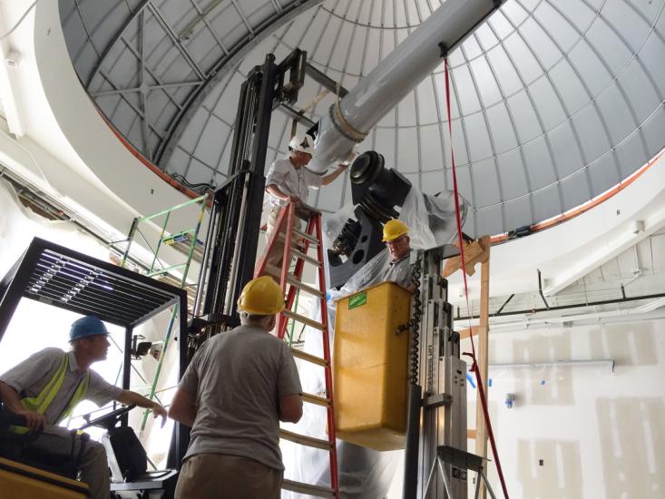 Large white telescope tube suspended from life as man on ladder guides it into place on a large base. Other staff are nearby on a bucket lift and on a forklift.