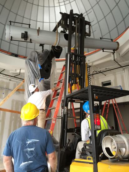 Large white telescope on heavy metal pier with man on ladder and others standing below as they  work on it.