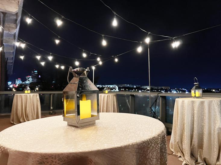 Terrace at night with skyline visible in background and string lights set up above three formally set tables