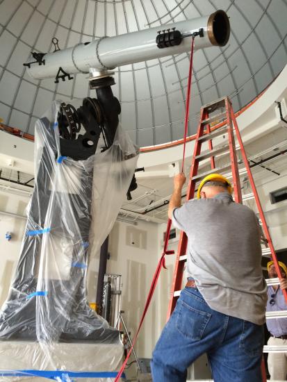 Man facing away from camera stands holding a rope attached to the cylinder of a large white telescope above him
