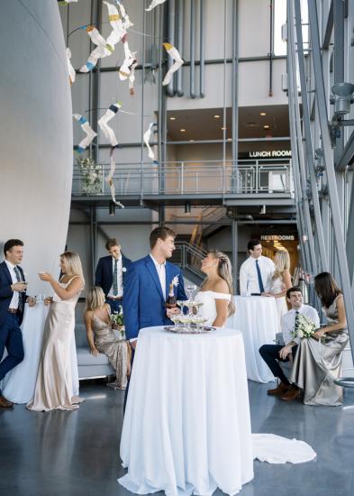 Bride and groom stand behind small, tall table with white tablecloth and members of their wedding party socializing in the background with the side of the planetarium dome exterior visible on the left side of the frame.
