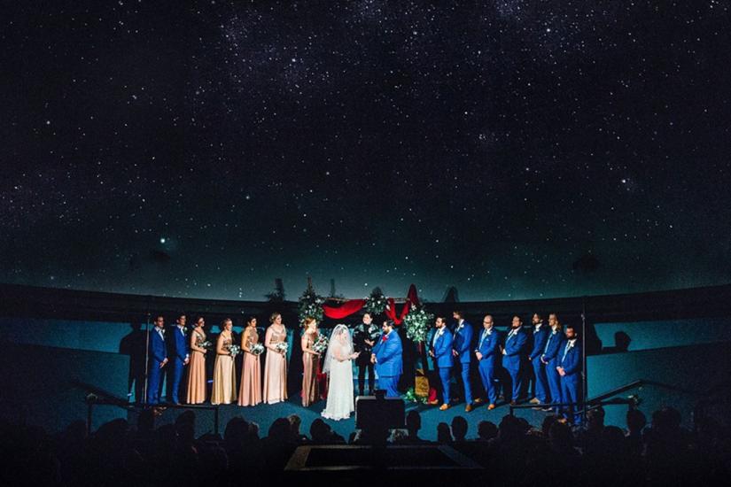 Wedding ceremony in the planetarium with couple flanked by bridesmaids and groomsmen with large projection of a starry night sky behind them.