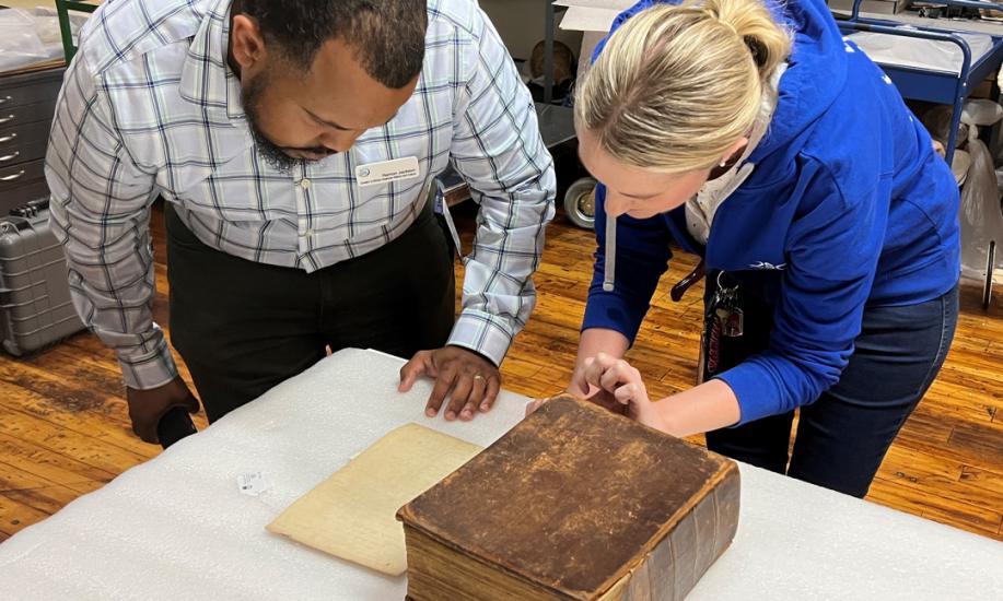 A man and woman bend down towards a table to examine a large, old leather bound book
