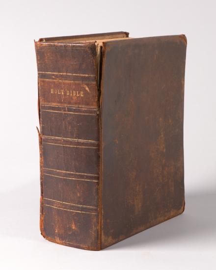 Large brown leather bound Bible stand vertically with spine facing camera and "Holy Bible" written on it.