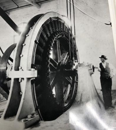 Black and white image of a man with a hat and pocket watch standing next to what looks like a very large piece wheel shaped machinery that is more than twice as tall as the man standing next to it.