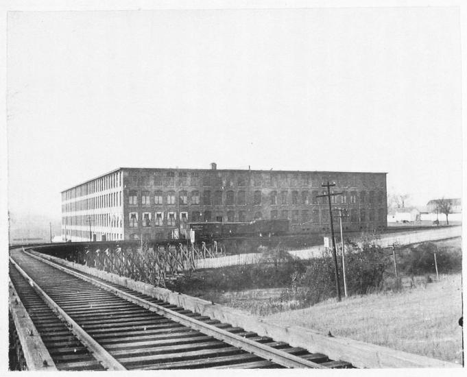 Black and white photograph shows train tracks in the foreground with a large brick, four story building in the background
