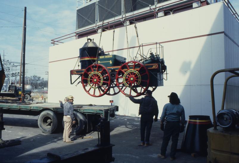 Life size antique train engine car is suspended on a lift as it is moved from the back of a flatbed truck to the ground.