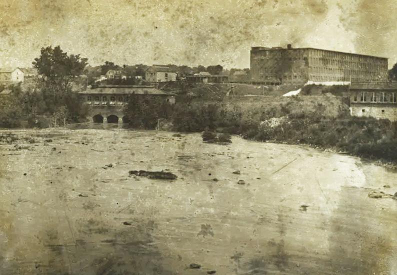 Old, sepia toned photograph shows canal in the foreground with large brick building on a rise above the canal