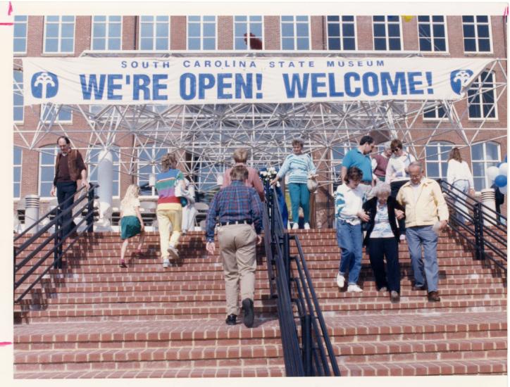 Group of people walking up stairs towards a large brick building with a banner over the entrance that reads "We're Open! Welcome!"