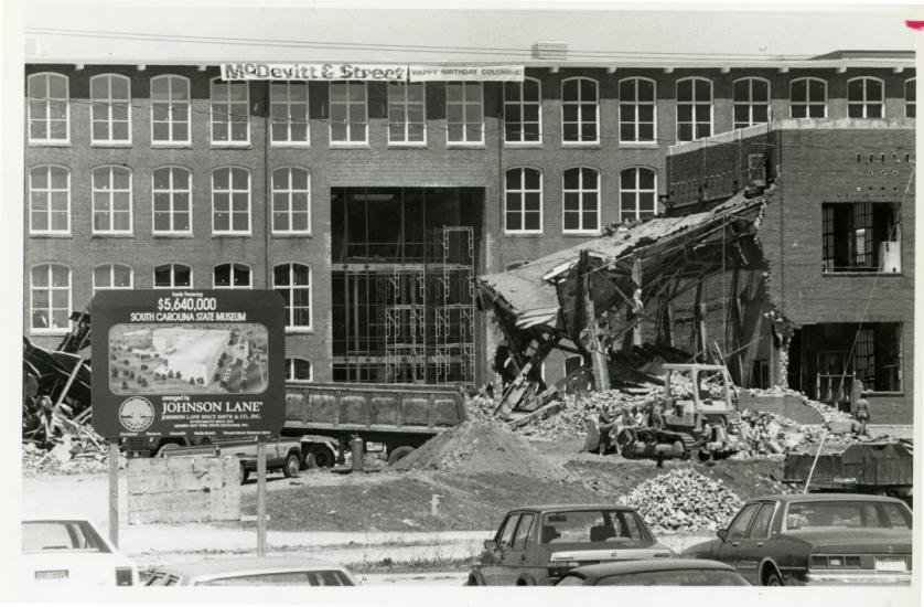 Black and white image of long, four story textile mill building under renovation. A sign is posted in front promoting the 5.6 million dollar project.