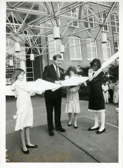 Black and white images shows 4 adults (1 man and 3 women) cutting through a large ribbon with an oversize pair of scissors.