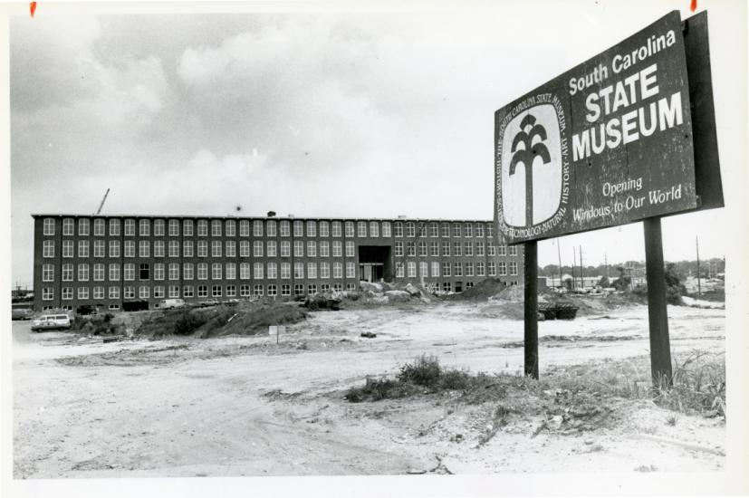 Black and white image of long, four story textile mill building under renovation. A sign is posted in front that reads "South Carolina State Museum Opening Windows to Our World".