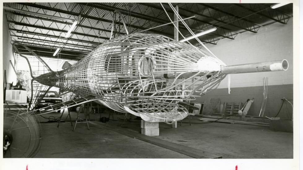 Black and white shows very large metal skeleton of a Megalodon shark in a workshop-like space