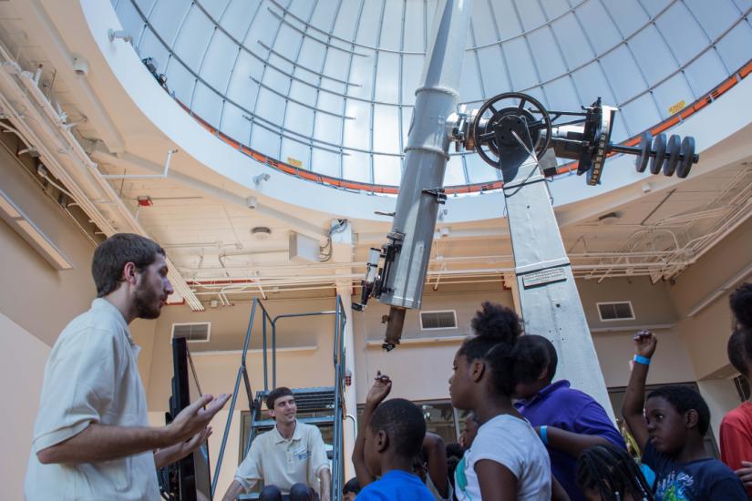 Bearded man on left side of the image addresses a group of young students with a huge telescope just behind them.