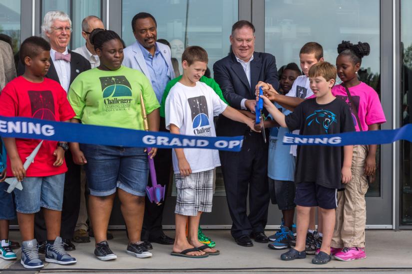 Group of older and younger people stand behind a blue ribbon that is being cut by a man holding an oversize pair of blue handled scissors.