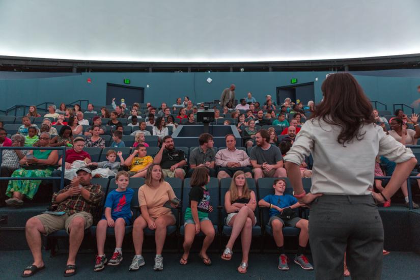 Woman with long dark hair stands with her back to the camera. An audience of people is seated in front of her in rows of blue chairs under a large, domed projector screen.