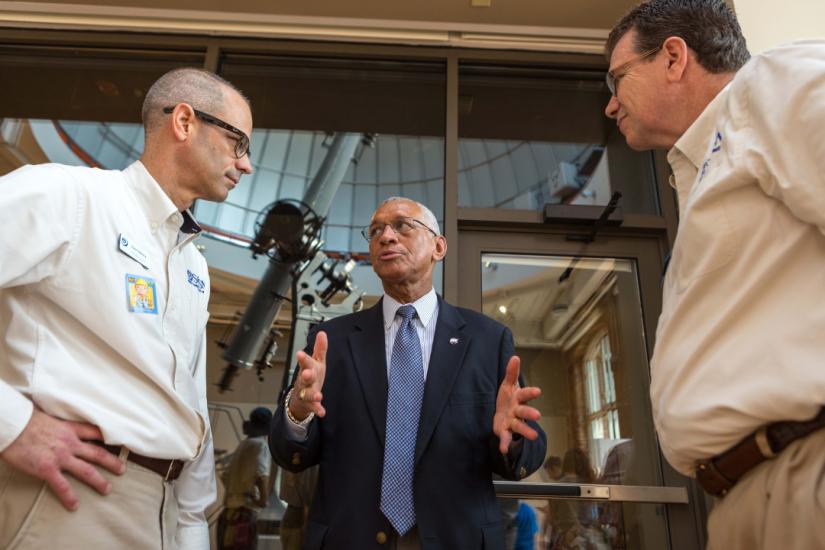 Two men in light colored button down shirts stand looking at another navvy blue suited man between them as that man gesticulates while speaking to them