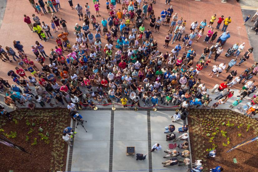 Image looks down from a large height onto a large group of people standing below, in front of the main entrance to a building.