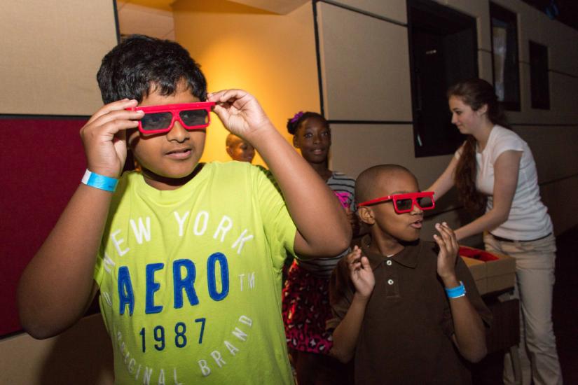 Young boy in yellow t-shirt with hands up to either side of his eyes as he adjusts a pair of red 3D glasses