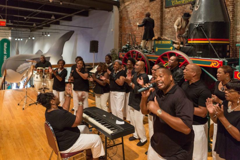 Group of singers, keyboard player and drummer in black shirts perform in front of a life size train replica