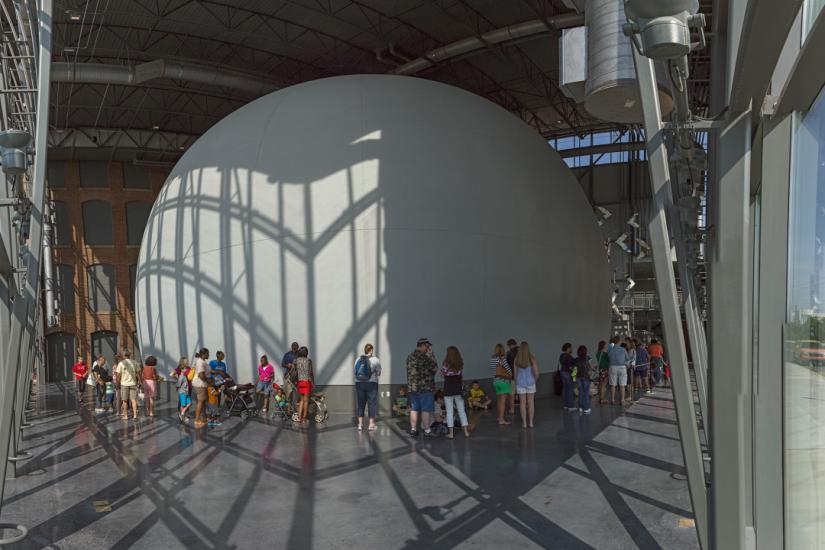 Large space with glass walls, a polished gray floor and a large gray dome structure with a line people standing in front of it