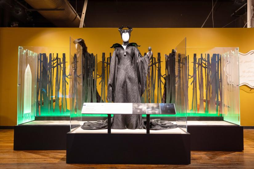 Platform with low panel in front. On the platform is a tall mannequin wearing a long and black gown with a large collar and a headdress featuring two curvy horns