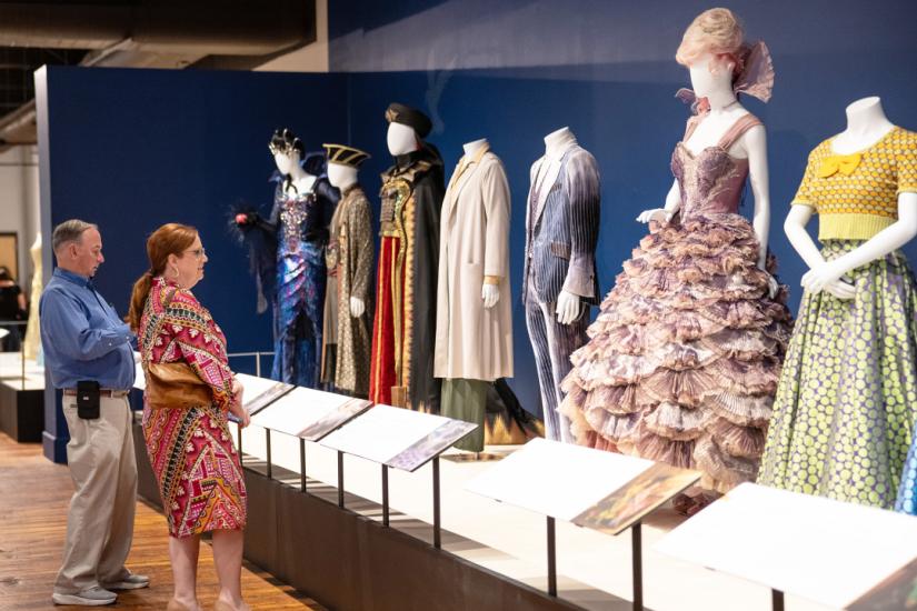 A man and woman stand looking up at a row of 7 mannequins that are dressed in various costumes, including a frilly lavender gown, a gray velvet suit, black and red robes with an embroidered cape.