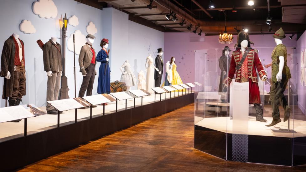 Room with hard wooden flooring and a row of platforms displaying 8 mannequins in various costumes, including weathered pants and brown jacket, a royal blue women's dress suit with red heeled shoes, a white slim feathered gown and more. Two mannequins on a platform to the rights side are wearing a pirate costume with a red velvet embroidered coat and bicorn hat and the other figure (Peter Pan) has forest green trousers boots, and a hat.