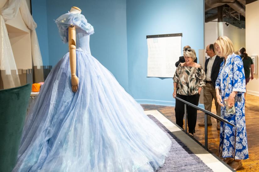 Side view of mannequin wearing voluminous pale blue ball gown with small butterfly appliques across the bodice. Two women stand on the right, one in a blue and white dress and the other in black pants and patterned blouse, stand looking at the ball gown.