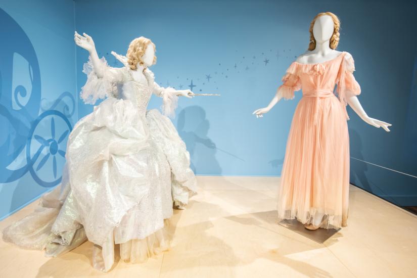 Two mannequins stand with one dressed in a voluminous light silvery white gown holding a thin wand pointed at the other figure which is wearing a tattered pale pink dress.