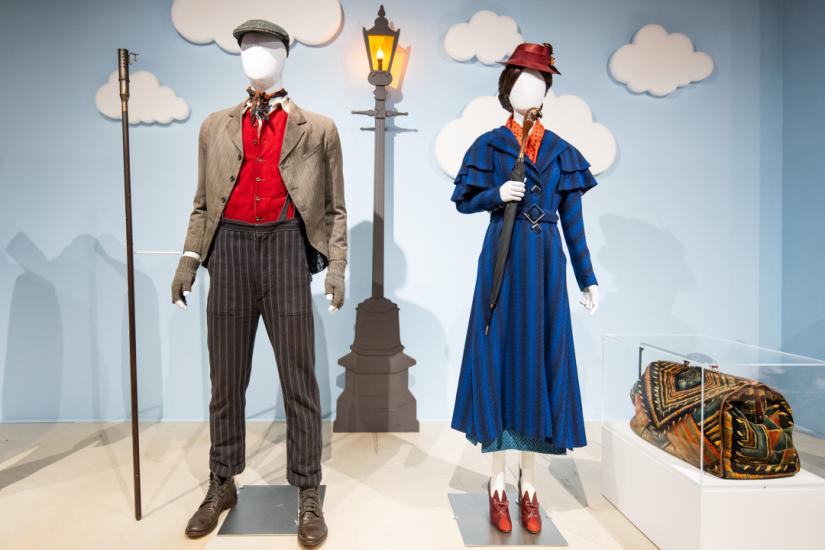 Two mannequins on plaform. The wall behind them in plae blue and decorate with clouds and the image of a light post lit with a gas flame. One figure is dress in brown striped trousers, a red vest and a tan dirty jacket. The other is dressed in a royal blue ladies' dress suit with red heeled shoes and a red hat. She is holding an umbrella with a handle carved like bird's head.