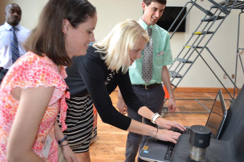 Two women look a computer screen while one types with man standing next to them in a green shirt and tie.