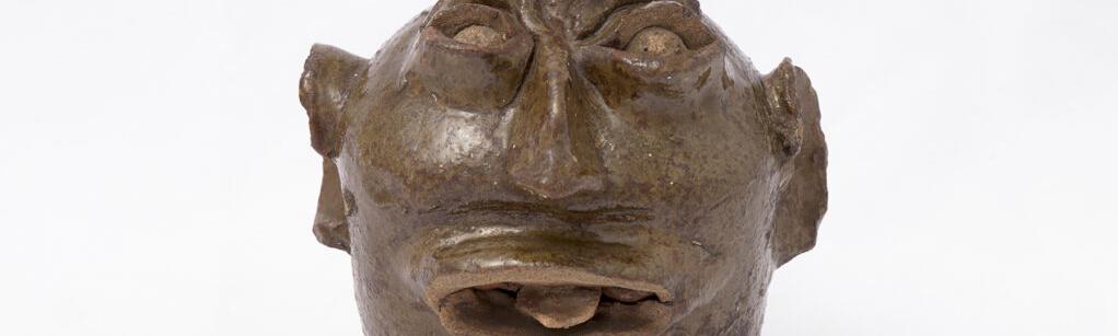 Small ceramic jug with odd looking face featuring large ears and nose, mouth open slightly with a protruding tongue.