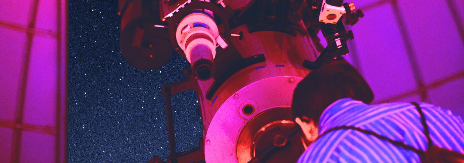 Telescope pointed at the night sky which is visible through an opening in a domed roof. The image is washed in reddish light.