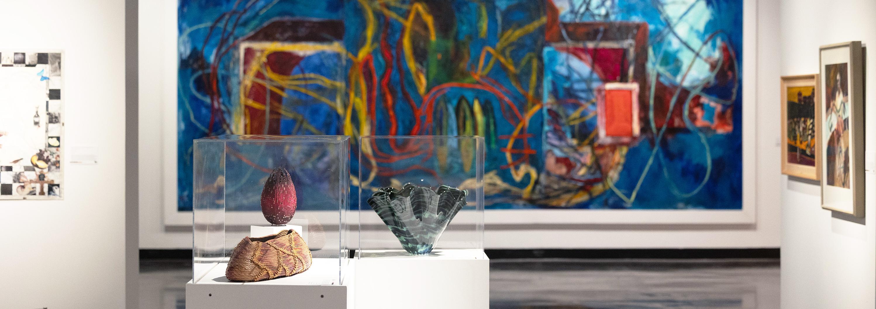 Large wall covered in colorful abstract paining in deep blues, yellow, red and purple. In the foreground is a small case with a beaded sculpture a large glass bowl on display.