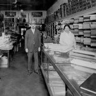 Black and white shows a man and woman standing in a shop with folded textiles in shelves on the walls.